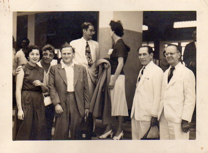A group of six people, dressed in 1940s attire, socialize indoors; a woman in a skirt stands beside a man while others conversate nearby.