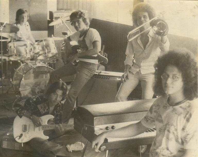 Five musicians in a vintage studio pose with instruments: drums, guitar, trombone, and keyboard. The setting reflects a retro musical vibe.