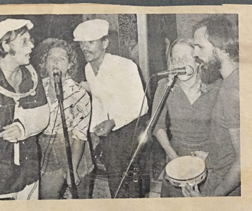 Group of five musicians performing together, singing into microphones. One is playing a tambourine, capturing a lively music scene.