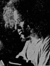 A musician with curly hair is focused on playing a guitar, illuminated by soft lighting that creates a moody atmosphere, emphasizing their concentration.