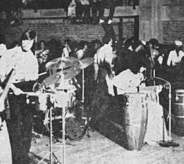 A lively band performs on stage, featuring a drummer and percussionist, in front of an enthusiastic crowd in a dimly lit venue.