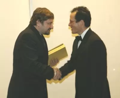 Two men in formal attire shake hands at an award ceremony, with one holding a yellow book, symbolizing recognition or achievement.