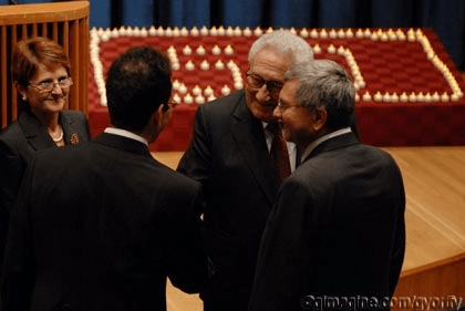Henry Kissinger engages in conversation with three individuals at a formal event, with lit candles in a backdrop. The setting suggests a significant gathering.